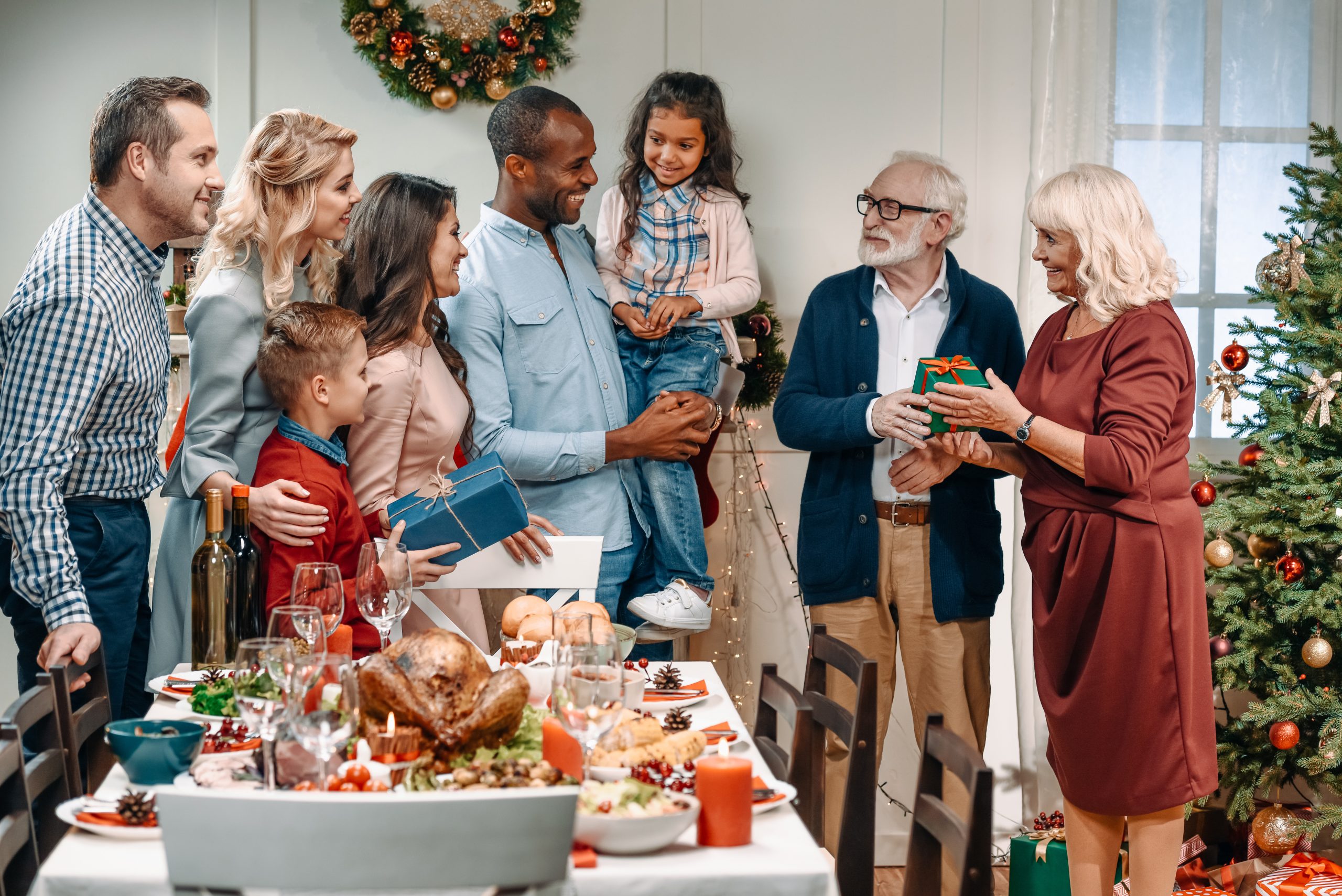 Multi-generational family gathered around a holiday dinner table exchanging gifts and smiling together.